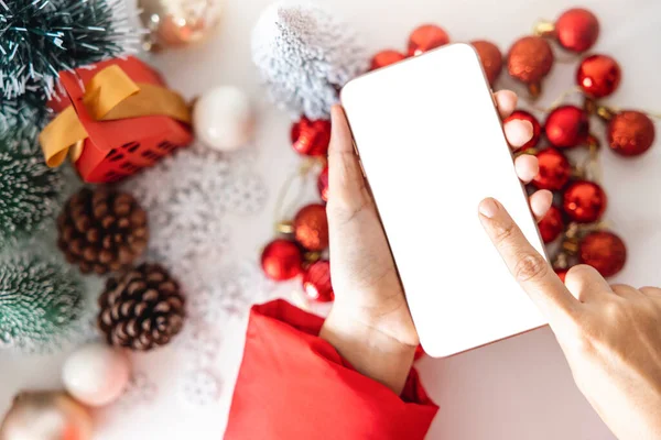 Top view Gentle hands cradle smartphone with a white screen mock and Fingers on the screen to shop online up by the festive scene of beautiful Christmas tree adorned, with Christmas balls, pine cones.