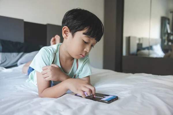 Close up portrait of Asian boy with black bangs, black eyes with a smiling face wearing a light green shirt lying on the bed in his house playing with his smart phone. Education concept.