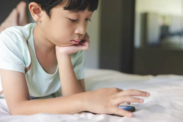 Close up portrait of Asian boy with black bangs, black eyes with a smiling face wearing a light green shirt lying on the bed in his house playing with his smart phone. Education concept.