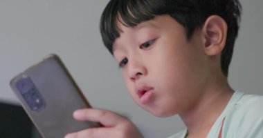 Siyah saçlı, gülümseyen yüzlü Asyalı bir çocuk evinde oturmuş akıllı telefonuyla oynuyor. Hayal gücünü kullanmaktan zevk alıyor. Çocuk kavramı, öğrenme, yaratıcılık, teknoloji, hobi. 4k