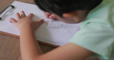 Siyah saçlı, gülümseyen yüzlü Asyalı bir çocuk evinin zeminine uzanmış örgülü bir kız resmi çiziyor. hayal gücünü kullanarak sanat yaratıyor. Çocuk kavramı, öğrenme, yaratıcılık. 4k