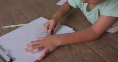 Siyah saçlı, gülümseyen yüzlü Asyalı bir çocuk evinin zeminine uzanmış örgülü bir kız resmi çiziyor. hayal gücünü kullanarak sanat yaratıyor. Çocuk kavramı, öğrenme, yaratıcılık. 4k