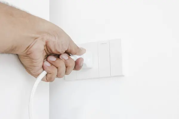 Hand plugging in an electrical appliance on white wall. Plug studio apartment power supply. Light switch. Power outlet. Concept of technology, connection, power saving, energy saving.