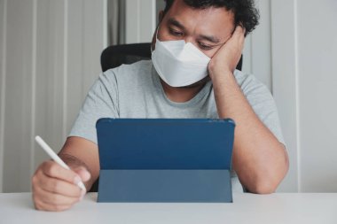 Evde yüz maskesi takan sıkkın adam salgın sırasında tablet ve stil ile çalışıyor, yorgun ve motivasyonsuz, uzaktan çalışma yorgunluğu, odaklanma eksikliği, salgın yorgunluğu, dijital yaşam tarzı.