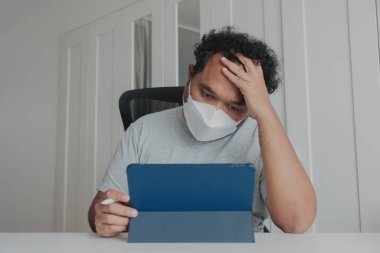 Evde yüz maskesi takan sıkkın adam salgın sırasında tablet ve stil ile çalışıyor, yorgun ve motivasyonsuz, uzaktan çalışma yorgunluğu, odaklanma eksikliği, salgın yorgunluğu, dijital yaşam tarzı.