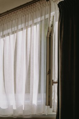 Açık bir penceredeki perdelerin arasından güneş ışığı süzülür, içini sıcak ve davetkar bir ışıltıyla aydınlatır..