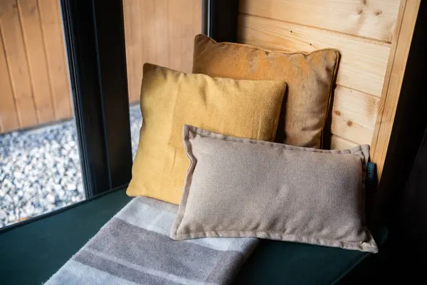 stock image A snug corner features three cushions of varying textures and a soft blanket on a dark green seat, against a backdrop of wooden walls and gray stones outside.