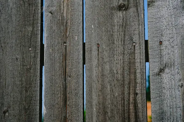 stock image A rustic wooden fence shows its age with worn planks and small gaps, framing a lush green background under clear blue skies.