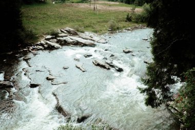 The clear river cascades over stones, surrounded by lush greenery and open fields, creating a calm and picturesque natural setting. clipart