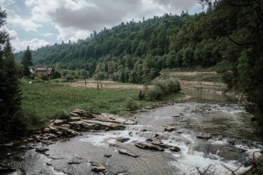 Bir nehir, uzak bir ev ve dağlık bir zemin eşliğinde, parçalı bulutlu bir gökyüzünün altında, yeşil bir vadide esiyor..