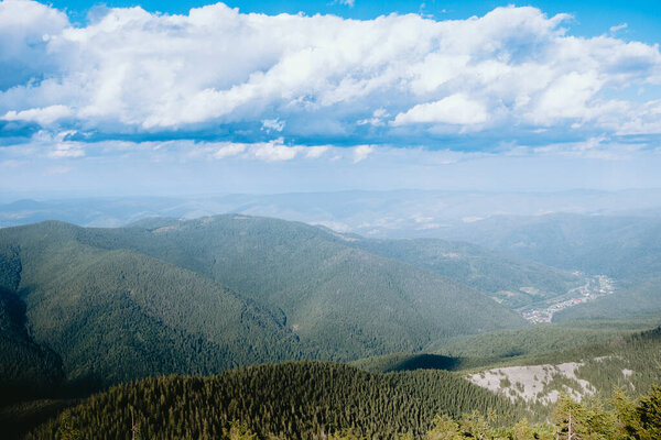 A sweeping view of lush green mountains and a valley below, with a small town visible in the distance, surrounded by impressive clouds.