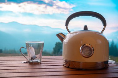 Çaydanlık boş bir cam bardağın yanındaki ahşap masada oturur, arkasında dağlar ve mavi bir gökyüzü vardır, bu da sakin bir sabah atmosferini gösterir..