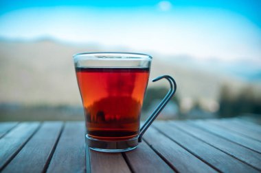 Dumanı tüten bir çay fincanı ahşap bir masanın üzerinde duruyor, uzak dağları ve arka planda parlak mavi gökyüzünü gösteriyor..