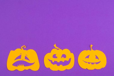 Üç parlak sarı balkabağı kesiği Cadılar Bayramı için mor bir zeminde neşeli yüzler sergiliyor..