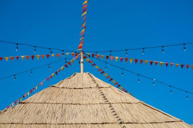 Bunting bayrakları ve sicim ışıkları sazdan çatı yapısını süsler, açık mavi gökyüzünün altında şenlikli bir atmosfer yaratır, kutlamalar veya açık hava toplantıları için mükemmeldir..