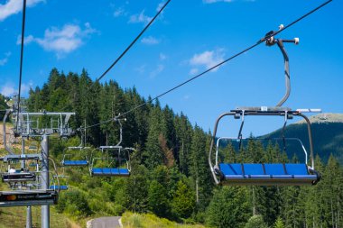 Binicisiz bir teleferik, sık orman ve açık gökyüzü tarafından çerçevelenmiş dağlık bir alanda yükselir ve yazın güzelliğini vurgular..