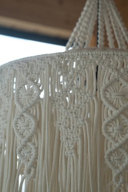 Çok güzel yapılmış bir makrome avize tavandan sarkıyor, ayrıntılı düğümler ve çıkıntılar sergiliyor, odayı doğal ışıkla aydınlatıyor..