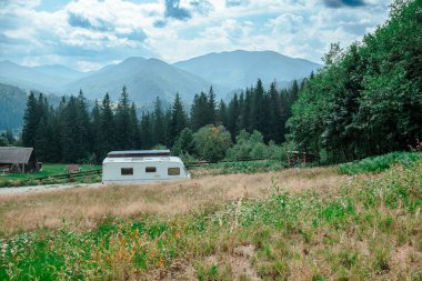 Eski bir karavan, parçalı bulutlu bir gökyüzünün altında uzun ağaçlar ve görkemli dağlarla çerçevelenmiş çimenli bir alanda dinlenir..