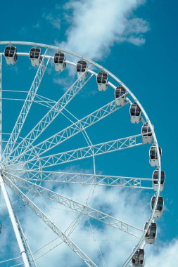 Dev dönme dolap dimdik ayakta duruyor, gondollarını pofuduk bulutlarla dolu canlı mavi gökyüzüne karşı sergiliyor..