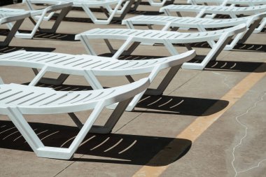 Güneş ışığı, havuzun yanındaki beyaz şezlonglara gölgeler düşürür. Rahatlamak için sakin ve davetkar bir ortam yaratır..