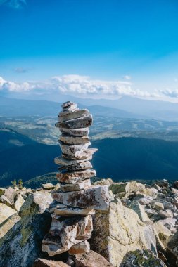 Stacked stones are carefully balanced on a mountain summit, overlooking a beautiful valley under a clear blue sky. The scene captures nature's tranquility. clipart