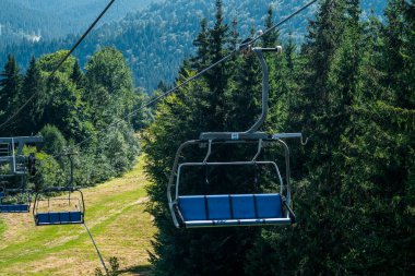 Bir teleferik, sakin bir yaz manzarasını gözler önüne seren dağların ve sık ağaçların ardında boş boş duruyor..
