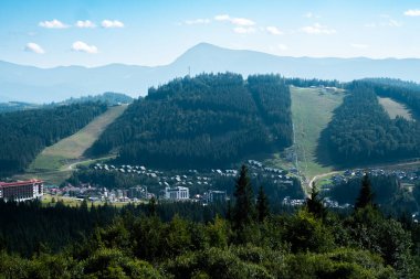 Geniş dağ manzarasında yemyeşil yamaçlar, bir kayak merkezi ve açık mavi gökyüzünün altında uzak bir tepe bulunur..