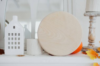 Şömine rafında beyaz bir ev, bir mum tutacağı ve sonbahar yapraklarının yanında yuvarlak bir tahta parçası sergileniyor..