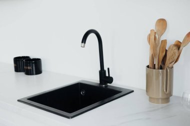 Gösterişli siyah bir lavabo şık bir muslukla eşleştirilirken ahşap çatal bıçaklar mermer tezgahın üzerindeki şık bir kabın içinde duruyor ve çağdaş bir mutfak atmosferi yaratıyor..