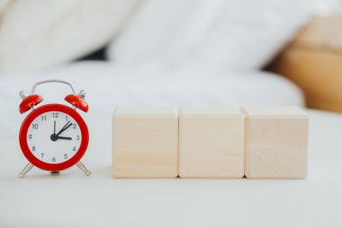 Kırmızı alarmlı bir saat zamanı gösterir. Bu sırada doğal ışık dolu, rahat bir yatak odasında, yakınlarda üç boş tahta blok bulunur..