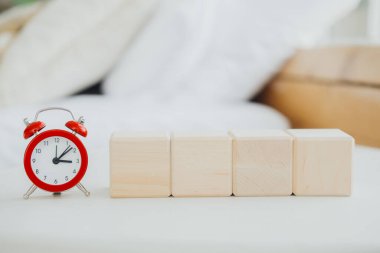 Bir çalar saat, yumuşak, beyaz bir yatak örtüsünün üzerindeki dört tahta bloğun yanında durur. Rahat ve davetkar bir yerde, bir sabah rutini ya da eğlenceli bir aktivite izlenimi uyandırır..