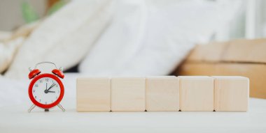 Sıcak ve davetkar bir oturma odasındaki bir masanın üzerinde düzgünce yerleştirilmiş beş tahta bloğun yanında kırmızı bir çalar saat duruyor. Yumuşak arkaplan uzayın gevşek atmosferine ekler.