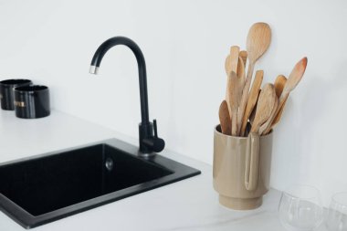 Çağdaş bir mutfakta, parlak siyah bir lavabo çeşitli ahşap mutfak aletleriyle dolu seramik bir tutacakla eşleştirilir ve şık ve işlevsel bir çalışma alanı oluşturulur..