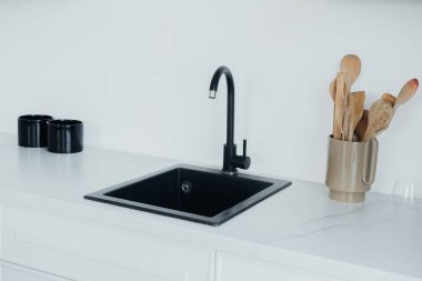 Pürüzsüz siyah bir mutfak lavabosu mermer bir tezgahın üzerinde, yanında da ahşap kaplar ve iki kahve fincanı bulunan seramik bir bardak ile minimalist ve şık bir yemek alanı oluşturur..