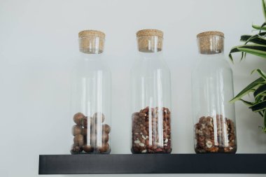 Mantar kapaklı üç cam kavanoz fındık ve tohumlardan oluşan bir çeşidi barındırıyor, siyah bir rafta ışık duvarına karşı zarif bir şekilde sergileniyor, uzaya doğal bir dekoru ekliyor..