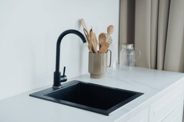 Parlak siyah bir lavabo beyaz bir tezgahın üzerinde duruyor, yanında da ahşap kaşıklarla dolu şık bir bej kap tutacağı var. Doğal ışık modern mutfak atmosferini geliştirir..