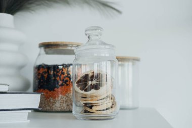 Renkli malzemeler ve kurutulmuş narenciye dilimleriyle dolu cam kavanozların yer aldığı şık bir düzenleme modern beyaz rafta çekici bir odak noktası oluşturuyor..