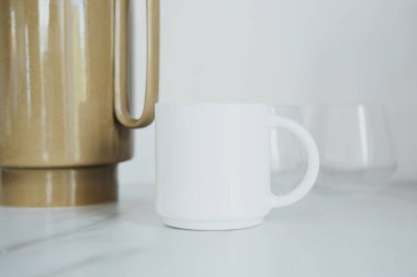 Sade beyaz bir kupa mermer bir tezgahın üzerinde parlak altın bir kahve demliğinin yanında oturur. Sahne, sıcak bir içecek için hazır minimalist bir mutfak öneriyor..