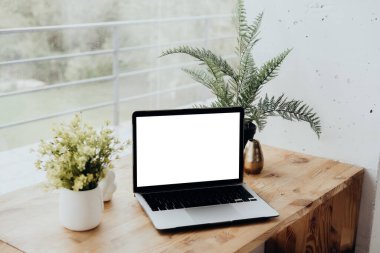 Bir dizüstü bilgisayar canlı yeşil bitkiler ve çiçeklerle birlikte ahşap bir masanın üzerinde duruyor. Doğal ışık yakındaki bir pencereden geçer ve sakin bir çalışma alanı atmosferi yaratır..