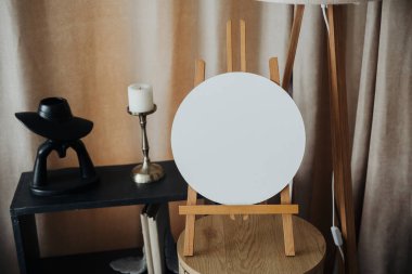 A wooden easel holds a blank round canvas, surrounded by a candle, a small black sculpture, and a stylish lamp in a warm, inviting room. The atmosphere hints at creativity and inspiration. clipart