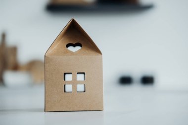 Kalp şeklinde kesim ve kare pencereli küçük bir mukavva ev temiz bir yüzeyde duruyor. Arka plan bulanık, bu büyüleyici dekorun ustalığını vurguluyor..