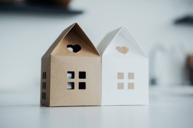 Açık kahverengi ve beyaz renkli iki dekoratif kağıt ev yan yana duruyor. Kesilmiş pencereler ve kalp şeklindeki üst açıklıklar modern iç dekorasyona eğlenceli bir dokunuş katıyor..