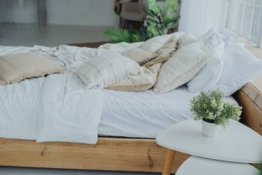 Konforlu bir yatak odasında yumuşak, katmanlı, nötr bir yatak sehpası, beyaz bir masa üzerinde küçük bir bitki, sakin ve davetkar bir atmosfer yaratıyor, rahatlamak için mükemmel..