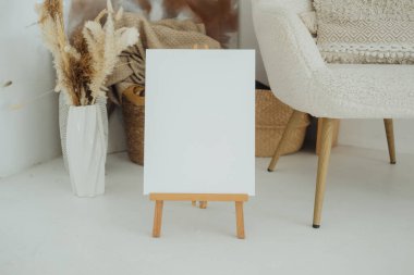 Boş bir tuval rahat bir sandalyenin yanındaki tahta bir sehpanın üzerinde duruyor. Dekoratif bitkiler ve desenli bir sepet sanat alanında sıcak bir atmosfer yaratarak yaratıcılığı davet ediyor..