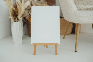 An empty easel is prominently displayed in a warm, inviting workspace, surrounded by plants and natural textures. The minimalist setting is perfect for creative inspiration. clipart