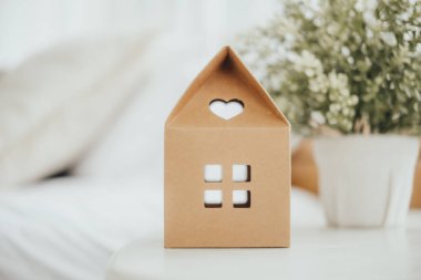 Kalp şeklinde kesiği olan büyüleyici bir mukavva ev, yumuşak ışıklandırma ve küçük bir saksı bitkisi ile çevrili bir masanın üzerinde oturur, rahat ve davetkar bir atmosfer yaratır..