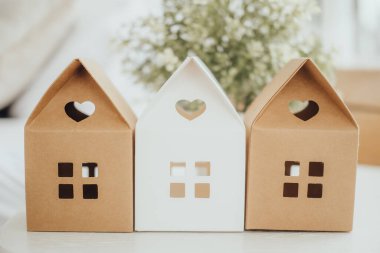 Three small paper houses in brown and white display heart-shaped cutouts. They add a whimsical touch to the cozy indoor setting, surrounded by soft light and greenery. clipart