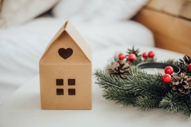 A charming cardboard house with a heart-shaped window sits next to a festive wreath adorned with red berries and pinecones on a cozy table, creating a warm holiday atmosphere. clipart