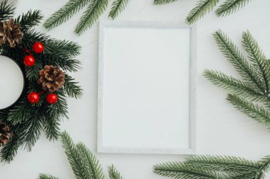 Merkezde boş bir çerçeve, taze çam dalları, kırmızı böğürtlenler ve kozalaklarla çevrili mevsimlik dekorasyon için ideal konforlu ve şen bir atmosfer yaratıyor..