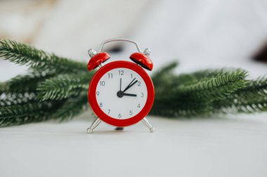 Canlı bir kırmızı çalar saat, tatil sezonunu sembolize eden yemyeşil çam dallarına dayanır. Arka plan sıcak ve davetkar bir atmosferin kış şenlikleri için mükemmel olduğunu gösteriyor..